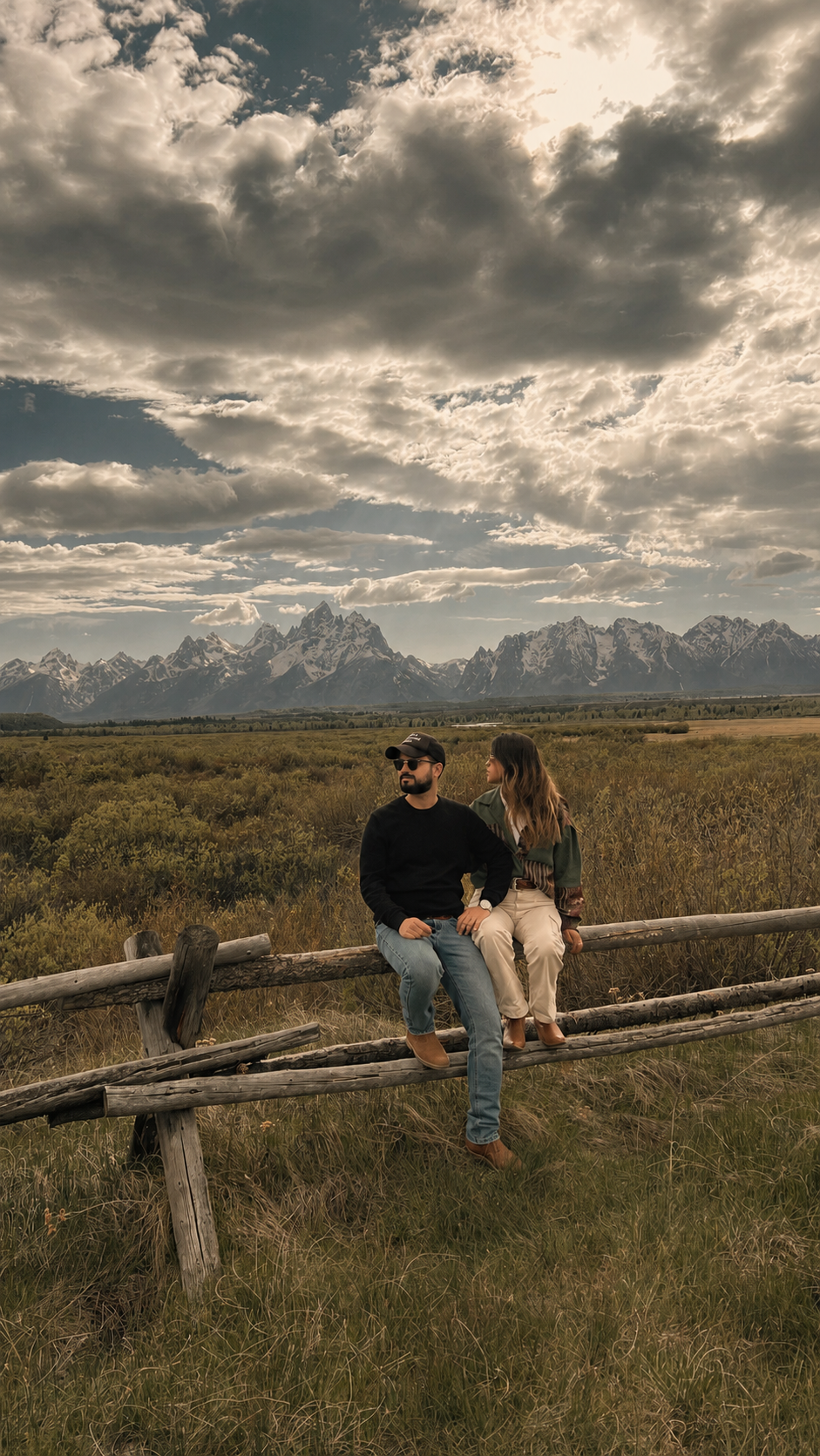 Casal sentado em uma cerca de madeira diante das montanhas Grand Teton